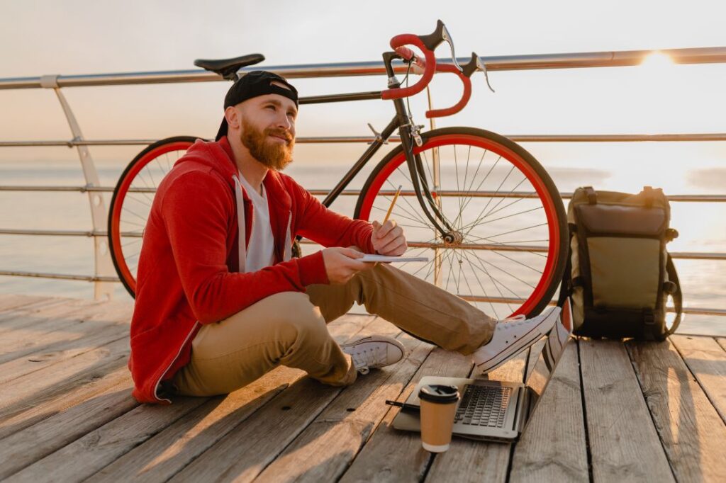 handsome hipster style bearded man working online freelancer laptop with backpack bicycle morning sunrise by sea healthy active lifestyle traveler backpacker