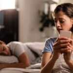 young woman is secretly sending text messages smart phone cheating her boyfriend who is sleeping bed