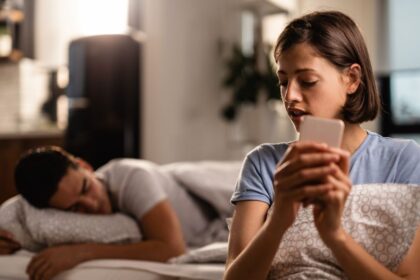 young woman is secretly sending text messages smart phone cheating her boyfriend who is sleeping bed