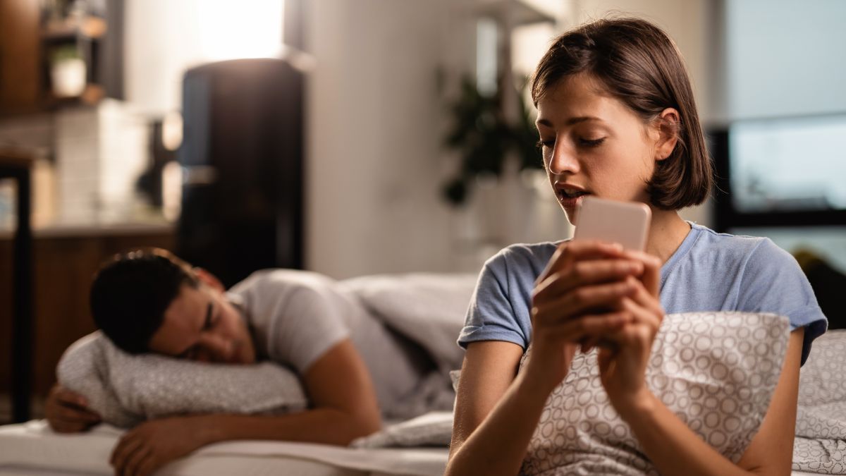 young woman is secretly sending text messages smart phone cheating her boyfriend who is sleeping bed